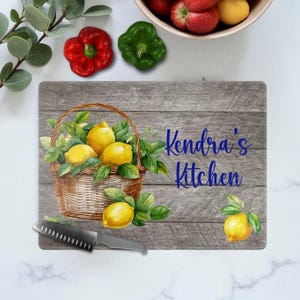 May include: Rectangular cutting board with a rustic wood grain design. It features a watercolor illustration of a basket filled with lemons and greenery. The text "Kendra's Kitchen" is written in blue script. A stainless steel knife rests on the board.