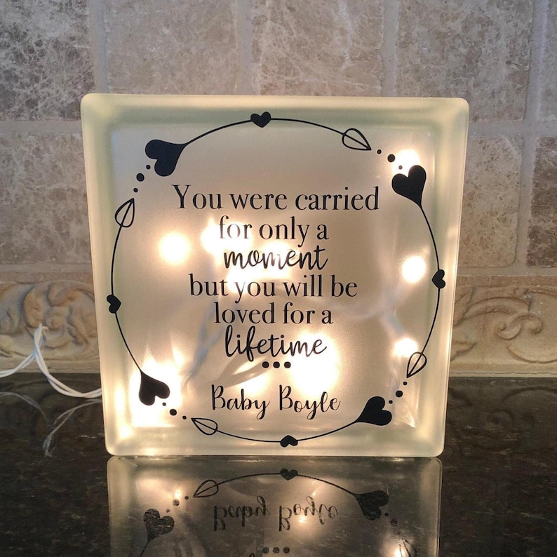 Puede incluir: Un bloque de vidrio esmerilado cuadrado con luces internas. El bloque presenta un dise&ntilde;o de coraz&oacute;n y hojas negras alrededor del texto: "You were carried for only a moment but you will be loved for a lifetime Baby Boyle."