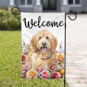 May include: Welcome garden flag featuring a golden doodle dog with a pink and yellow floral design. The flag has the word "Welcome" printed in black.