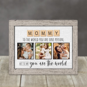 May include: A framed photo display with a light gray wooden frame. The display features the word "MOMMY" spelled out with Scrabble tiles above three family photos. The text "TO THE WORLD YOU ARE ONE PERSON, BUT TO ME you are the world" is also included.