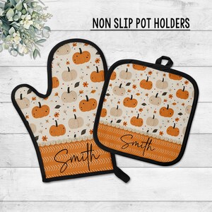 May include: A non-slip pot holder and oven mitt set. The set features a cream-colored background with orange and cream pumpkins, fall leaves, and the name "Smith" in script. The bottom border is orange with a chevron pattern.