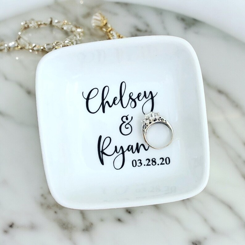 K&ouml;nnte beinhalten: Ein wei&szlig;es Keramik-Ringtablett mit abgerundeten Ecken. Auf dem Tablett stehen die Namen "Chelsey & Ryan" in schwarzer Schrift und das Datum "03.28.20". Ein silberner Ring mit einem gro&szlig;en Mittelstein liegt auf dem Tablett.