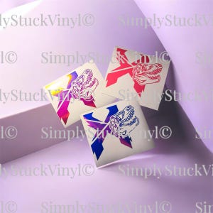 May include: Three square vinyl decals featuring a stylized dinosaur skull graphic overlaid on a geometric X shape. One decal has a red design, one has a blue design, and one has a multi-colored gradient design. Each decal is on a white backing.