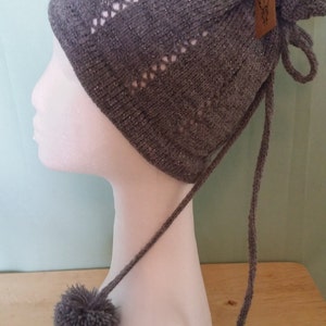 May include: Grey knitted hat with a long tie and two pom-poms. The hat has a delicate openwork pattern and a small tag attached to the tie.