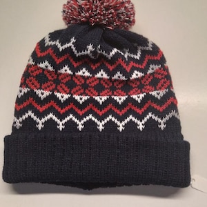 May include: A navy blue knitted beanie with a white and red geometric pattern. The beanie has a fluffy pom pom on top.