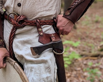 May include: A person wearing a tan linen shirt, leather shoulder armor, and arm bracers. A brown leather belt with an axe and a round shield are also visible. The person is standing in a forest setting.
