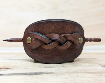 May include: A brown leather hair barrette with a braided design across the front. The barrette is secured with a wooden stick that is held in place by two brass rivets. The hair accessory is designed to hold hair in place.