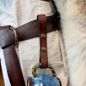 May include: A handmade blue ceramic mug with a twisted handle, suspended from a brown leather strap with brass rivets. The mug has a glossy finish with shades of blue and brown. The strap is attached to a brown leather belt.