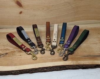 May include: A collection of leather wrist straps in various colors, including red, black, brown, cream, blue, purple, and green. Each strap features a metal clasp and a small metal plate with engraved text. The straps are displayed on a wooden surface.