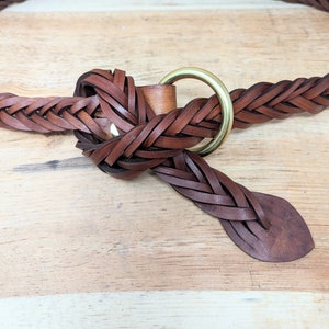 K&ouml;nnte beinhalten: Geflochtener brauner Lederg&uuml;rtel mit einer Messingringschlie&szlig;e. Der G&uuml;rtel besteht aus geflochtenen Lederstreifen und hat ein klassisches Design. Der G&uuml;rtel wird auf einer hellen Holzoberfl&auml;che gezeigt.