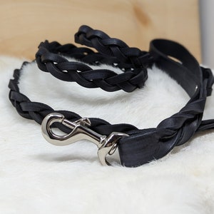 May include: A black leather dog leash and matching collar. The leash and collar are made of braided leather and feature a silver-colored metal clasp. The items are displayed on a white, fluffy surface.