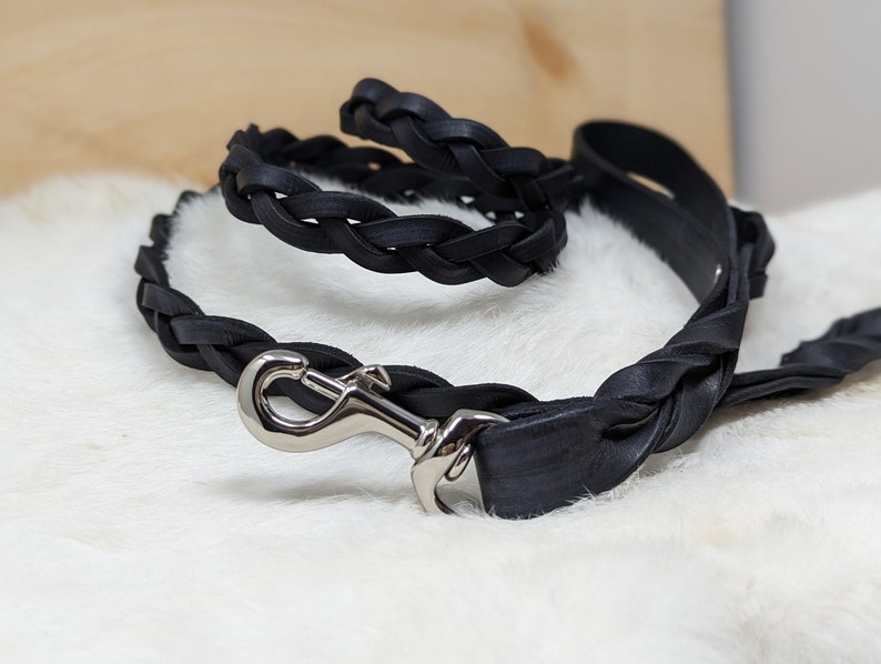 May include: A black leather dog leash and matching collar. The leash and collar are made of braided leather and feature a silver-colored metal clasp. The items are displayed on a white, fluffy surface.