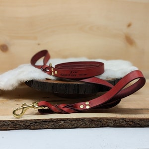 May include: A red leather dog leash with a braided handle and brass hardware. The leash is engraved with the name "Leo" and a phone number. The leash is coiled on a wooden surface.