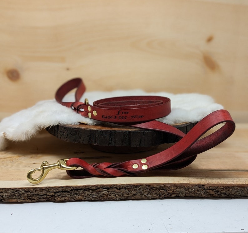 May include: A red leather dog leash with a braided handle and brass hardware. The leash is engraved with the name "Leo" and a phone number. The leash is coiled on a wooden surface.