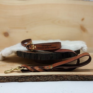 May include: A brown leather dog leash and matching collar set. The collar is engraved with the name "Cooper" and a phone number. The leash features a braided handle and a brass-colored clasp.