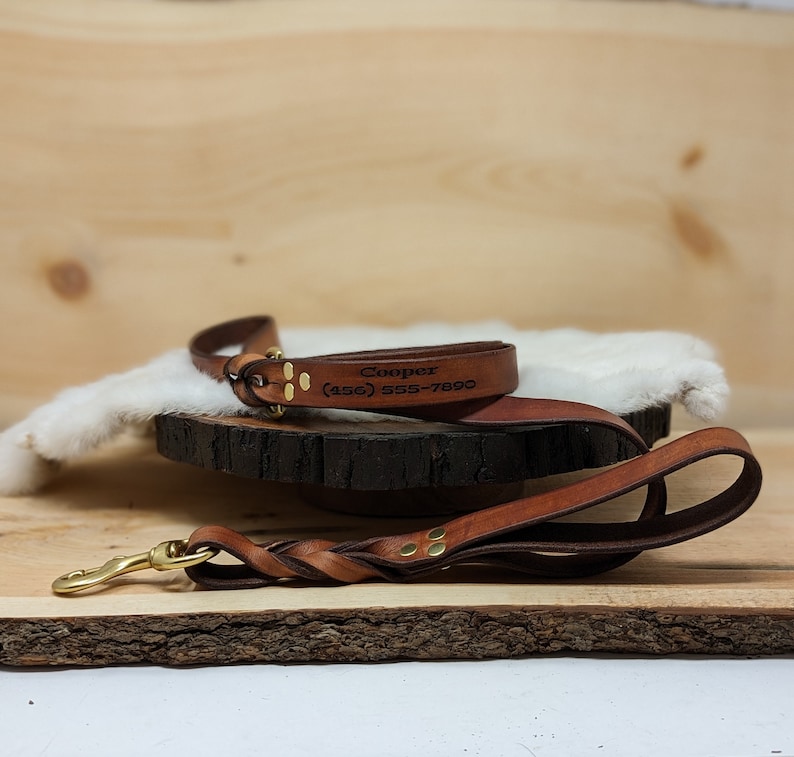 May include: A brown leather dog leash and matching collar set. The collar is engraved with the name "Cooper" and a phone number. The leash features a braided handle and a brass-colored clasp.