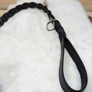 May include: A black leather dog leash with a braided handle and a silver-colored metal ring. The leash is shown against a white, fluffy background, with a wooden surface visible in the background.