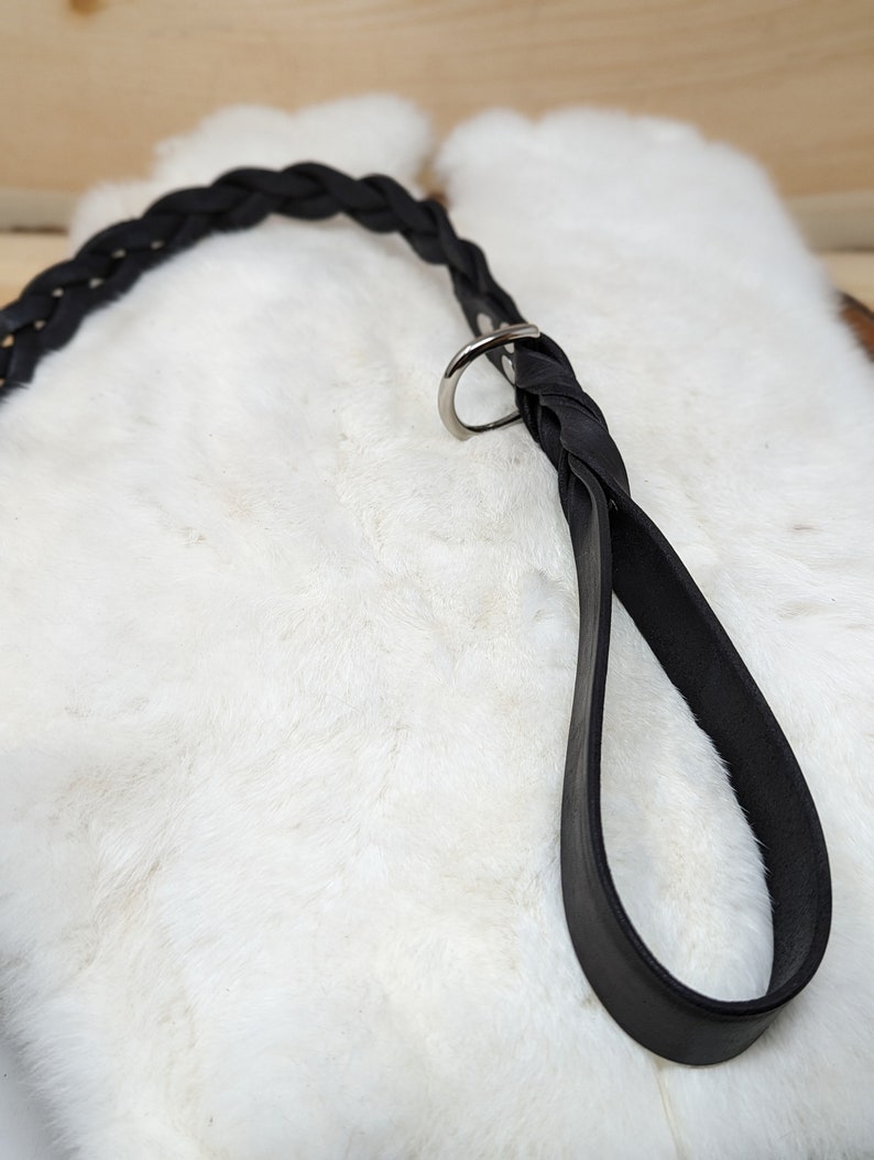 May include: A black leather dog leash with a braided handle and a silver-colored metal ring. The leash is shown against a white, fluffy background, with a wooden surface visible in the background.