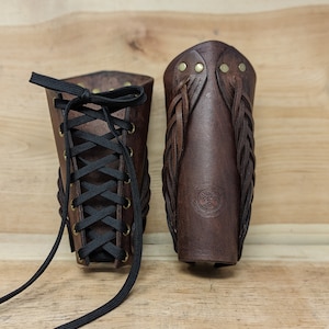 May include: Two brown leather arm bracers. One has a black lace-up closure, the other features braided leather accents and brass rivets. The bracers are displayed on a wooden surface, showcasing their craftsmanship and design.