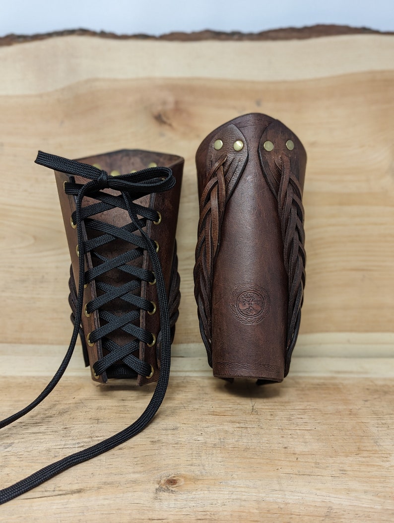 May include: Two brown leather arm bracers. One has a black lace-up closure, the other features braided leather accents and brass rivets. The bracers are displayed on a wooden surface, showcasing their craftsmanship and design.