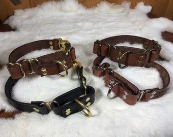 May include: Four leather dog collars in shades of brown and black, featuring brass hardware. The collars have a classic design with metal D-rings and adjustable buckles. The collars are displayed on a white, fluffy surface.