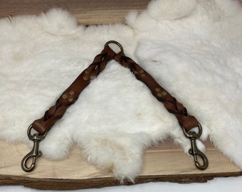 May include: Braided brown leather dog leash with brass-colored swivel clips and rivets. The leash is designed with a split end for attaching to two points on a dog's harness or collar. The leash is shown on a white fur surface.
