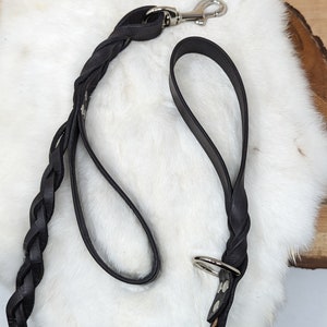 May include: A black leather dog leash with a braided design. The leash features a silver-colored metal clasp and a handle. The leash is lying on a white, fluffy surface.