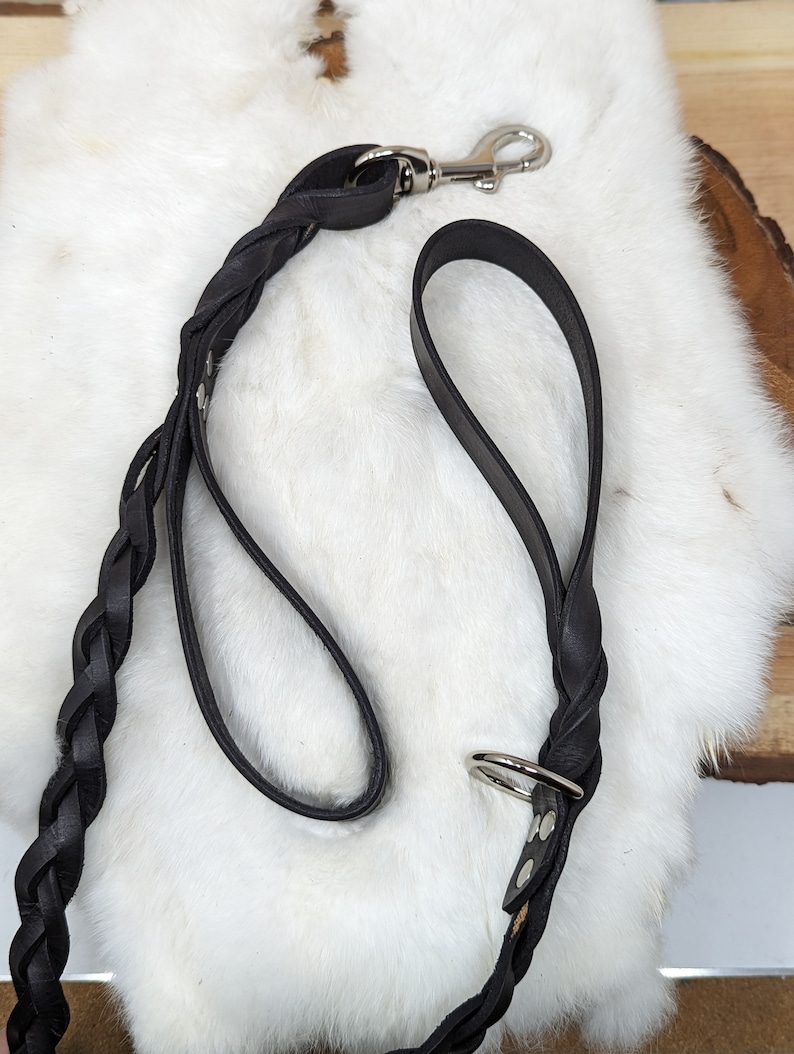 May include: A black leather dog leash with a braided design. The leash features a silver-colored metal clasp and a handle. The leash is lying on a white, fluffy surface.