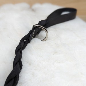 May include: A black leather dog leash with a braided design. The leash features a silver metal ring and a loop handle. The leash is lying on a white, fluffy surface.