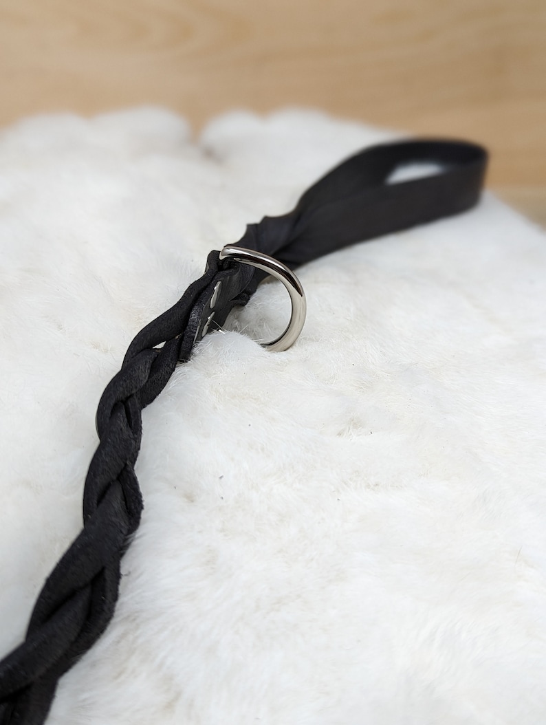 May include: A black leather dog leash with a braided design. The leash features a silver metal ring and a loop handle. The leash is lying on a white, fluffy surface.