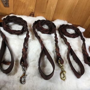 May include: Three brown leather dog leashes with braided sections and loop handles. Each leash has a metal clasp for attaching to a collar. The leashes are displayed on a white, textured surface.