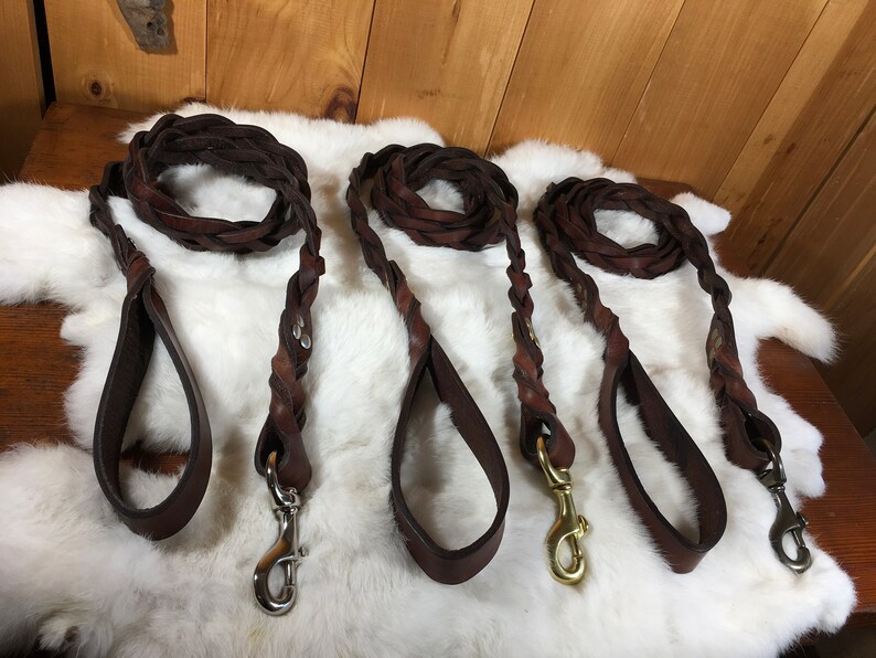 May include: Three brown leather dog leashes with braided sections and loop handles. Each leash has a metal clasp for attaching to a collar. The leashes are displayed on a white, textured surface.