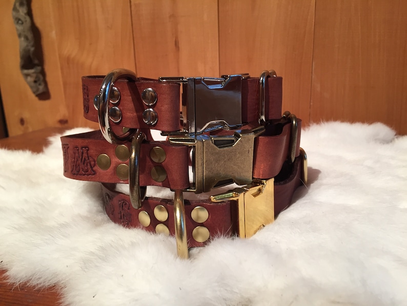 May include: Three stacked brown leather dog collars with metal hardware. The collars have engraved text, brass-colored rivets, and silver and gold-colored buckles. The collars are displayed on a white, fluffy surface.
