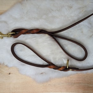 May include: A brown leather dog leash with a braided handle and a brass-colored clasp. The leash is lying on a white, fluffy surface, with a wooden background visible.