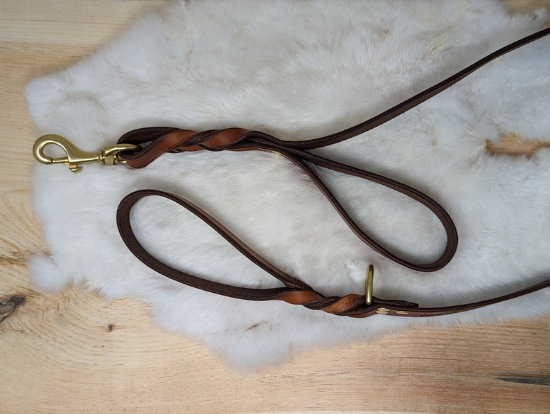 May include: A brown leather dog leash with a braided handle and a brass-colored clasp. The leash is lying on a white, fluffy surface, with a wooden background visible.