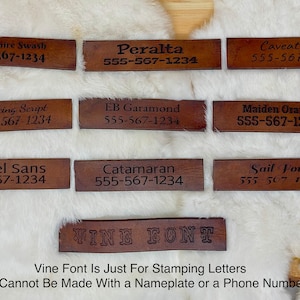 May include: Nine rectangular, brown leather tags with various font styles and phone numbers. The text includes "Berkshire Swash," "Peralta," "Caveat Font," "Dancing Script," "EB Garamond," "Maiden Orange," "Martel Sans," "Catamaran," and "Sail Font." A tenth tag reads "VINE FONT."