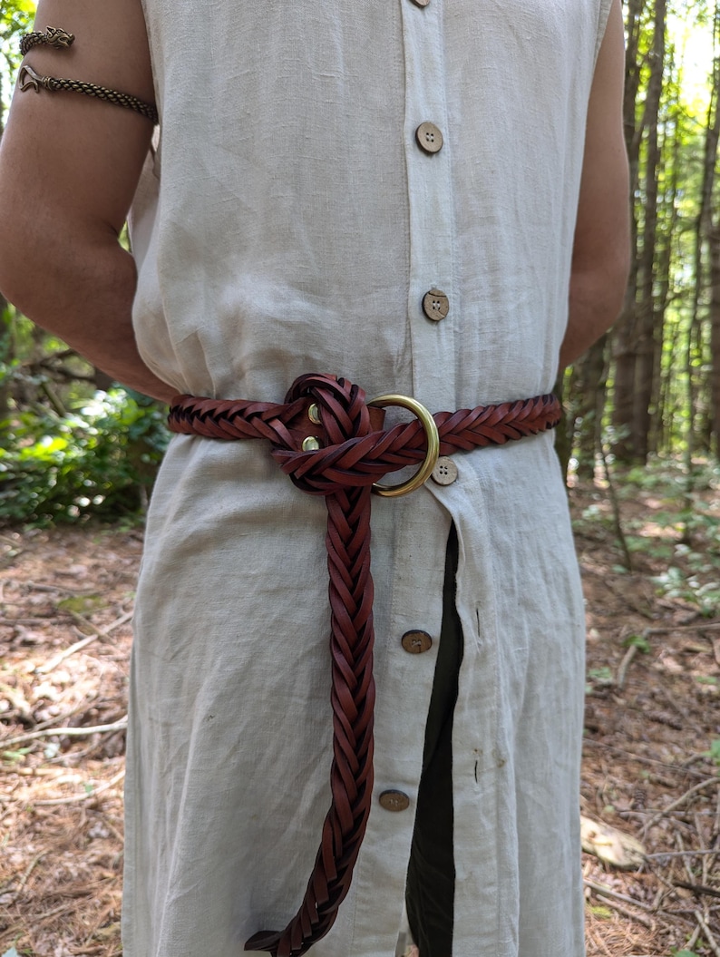 K&ouml;nnte beinhalten: Geflochtener brauner Lederg&uuml;rtel mit Messingringschlie&szlig;e. Der G&uuml;rtel wird &uuml;ber einem hellen Leinenkleid getragen. Ein passender Armreif ist am linken Arm zu sehen. Das Design des G&uuml;rtels weist ein geflochtenes Muster und ein Knotendetail auf.