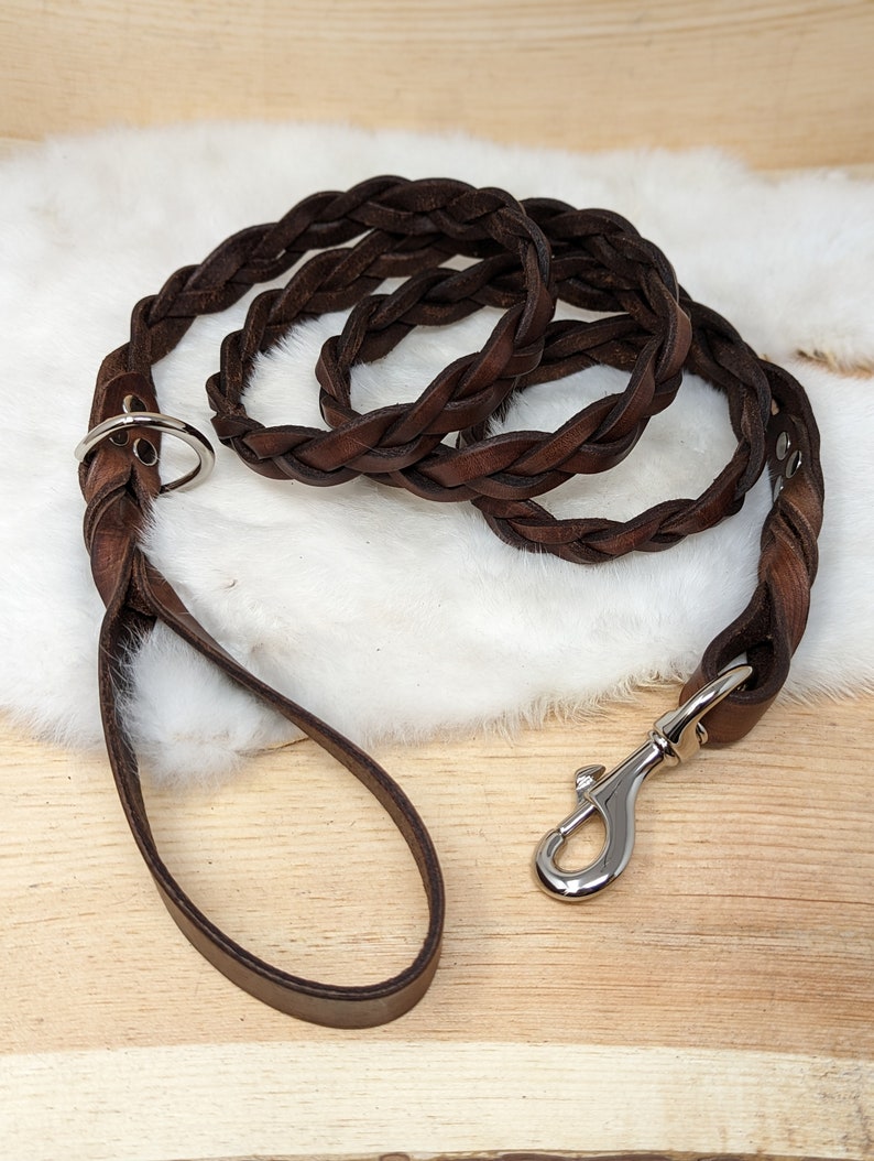 May include: Braided brown leather dog leash with a silver-colored metal clasp and ring. The leash is made of tightly woven leather strands, creating a durable and stylish design. The handle is a simple loop for easy gripping.