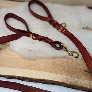 May include: Burgundy leather dog leash with a braided handle and brass hardware. The leash has the name "Leo" and a phone number engraved on it. The leash is lying on a wooden surface.