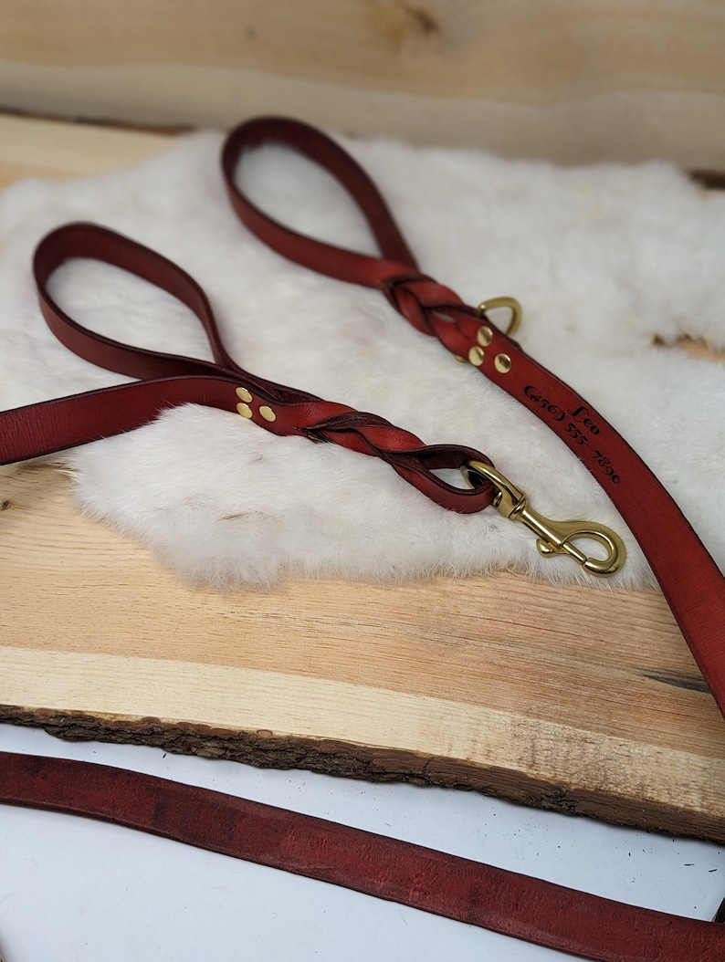 May include: Burgundy leather dog leash with a braided handle and brass hardware. The leash has the name "Leo" and a phone number engraved on it. The leash is lying on a wooden surface.