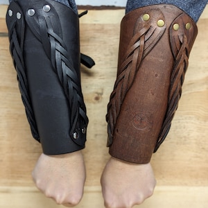 May include: Two leather arm bracers, one black and one brown, with braided leather accents. The bracers are secured with rivets and feature a decorative design. The brown bracer has a circular embossed emblem.