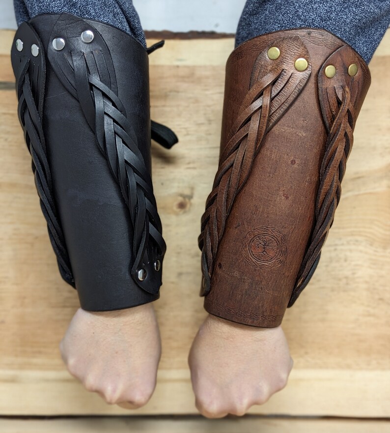May include: Two leather arm bracers, one black and one brown, with braided leather accents. The bracers are secured with rivets and feature a decorative design. The brown bracer has a circular embossed emblem.