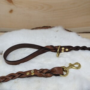 May include: Braided brown leather dog leash and matching handle with brass hardware. The leash features a brass clasp and is displayed on a white fur-like surface. The handle has a brass ring.