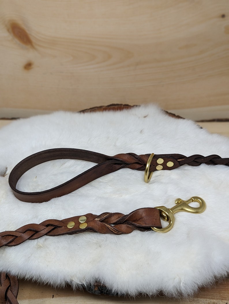 May include: Braided brown leather dog leash and matching handle with brass hardware. The leash features a brass clasp and is displayed on a white fur-like surface. The handle has a brass ring.