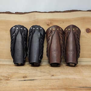May include: Four leather arm guards in black and brown, displayed on a wooden surface. The arm guards feature braided leather accents and are secured with rivets or studs. Two are black, and two are brown.