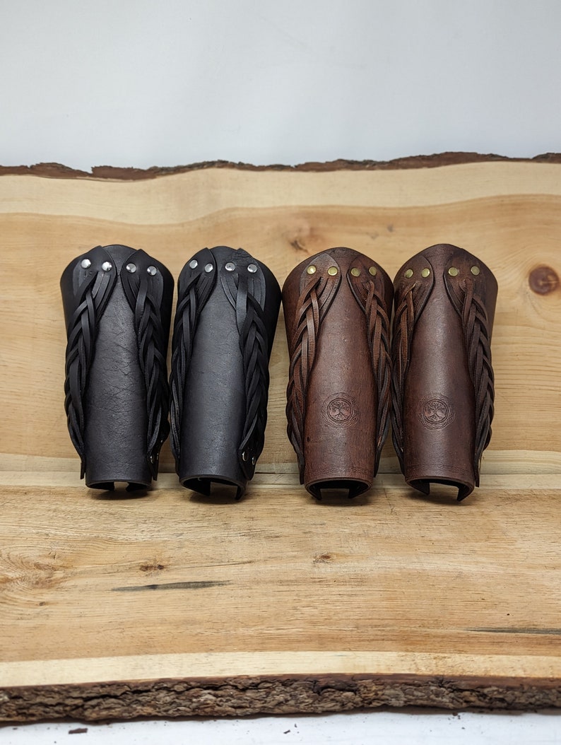 May include: Four leather arm guards in black and brown, displayed on a wooden surface. The arm guards feature braided leather accents and are secured with rivets or studs. Two are black, and two are brown.