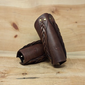 May include: Two brown leather arm guards with braided accents and brass rivets. The arm guards are cylindrical with a curved shape and a stamped emblem. They are displayed on a wooden surface.