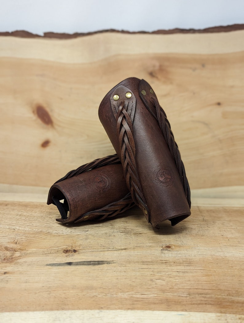 May include: Two brown leather arm guards with braided accents and brass rivets. The arm guards are cylindrical with a curved shape and a stamped emblem. They are displayed on a wooden surface.