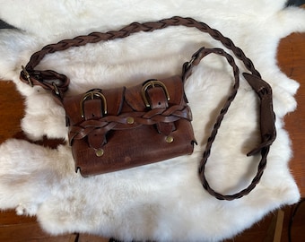 May include: A brown leather crossbody bag with a braided strap and brass buckles. The bag features a flap closure with two buckles and decorative braided leather accents. The bag is approximately 8 inches wide and 6 inches tall.