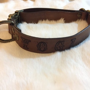 May include: A brown leather dog collar with a metal buckle and a large metal ring. The collar has the word "YOGI" embossed on it. The collar is designed to fit around a dog's neck.